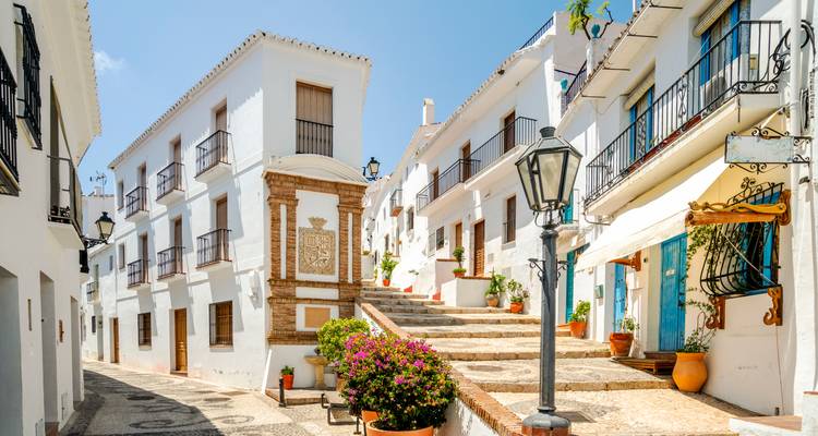 Heller andalusischer Dorfplatz mit weiß getünchten Häusern, Blumenkübeln und einer Treppe, die nach oben führt.
