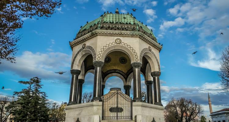 La ornada Fuente Alemana se alza majestuosa contra un cielo azul dramático con nubes, enmarcada por árboles invernales y pájaros que vuelan alto.