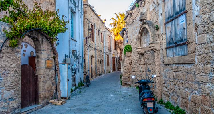 Narrow cobblestone alley with ancient stone walls, archways, and a parked scooter