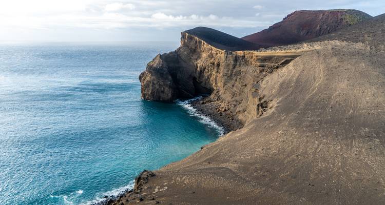 Promontoire volcanique spectaculaire de cendres noires et de rochers rencontrant les eaux océaniques turquoise sous un ciel nuageux.