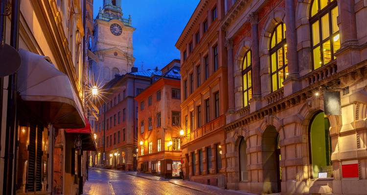 Kopfsteinpflasterstraße in Stockholms Altstadt bei Einbruch der Dunkelheit, beleuchtet von warmem Laternenlichter mit Kirchturm darüber.