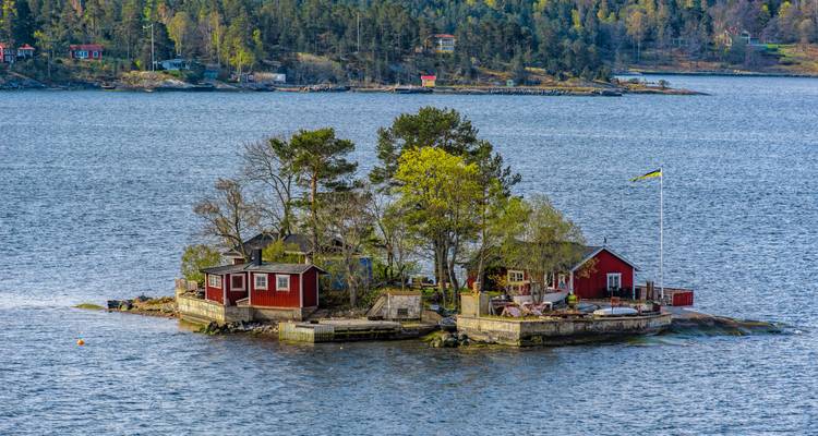 Winzige bewaldete Insel im Stockholmer Schärengarten mit roten Häuschen und einer schwedischen Flagge.