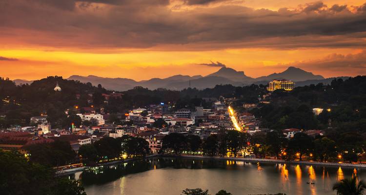 Die Lichter der Stadt Kandy funkeln um den See unter dramatischen orangefarbenen Sonnenuntergangswolken und Bergen
