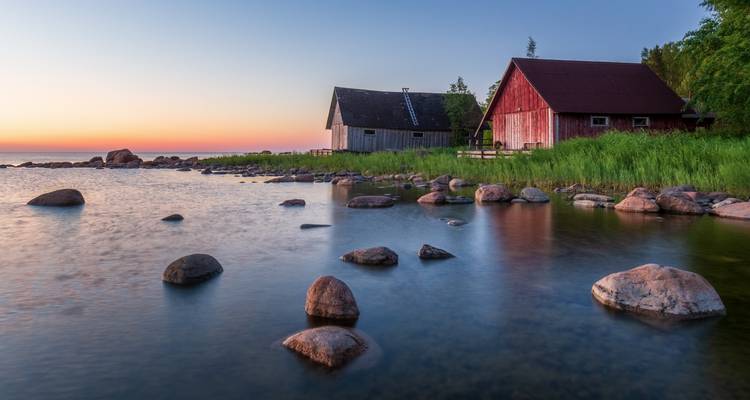 Ruhige Küstenszene bei Sonnenuntergang mit rustikalen Holzscheunen, glatten Steinen und ruhigem Wasser, das pastellfarbene Himmel spiegelt.