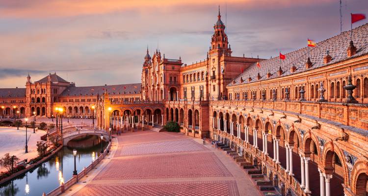 Plaza de España in Sevilla leuchtend in warmen Sonnenuntergangstönen mit Lichtern, die sich auf dem gemusterten Platz und dem Kanal widerspiegeln.