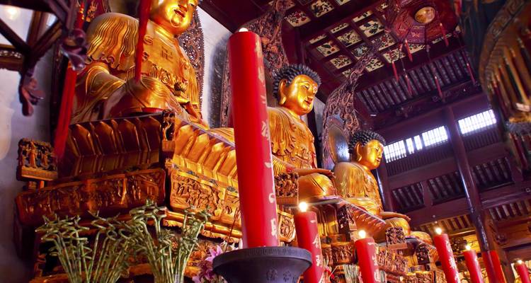 Goldene Buddha-Statuen und hohe rote Kerzen in einer kunstvoll verzierten hölzernen Tempelhalle