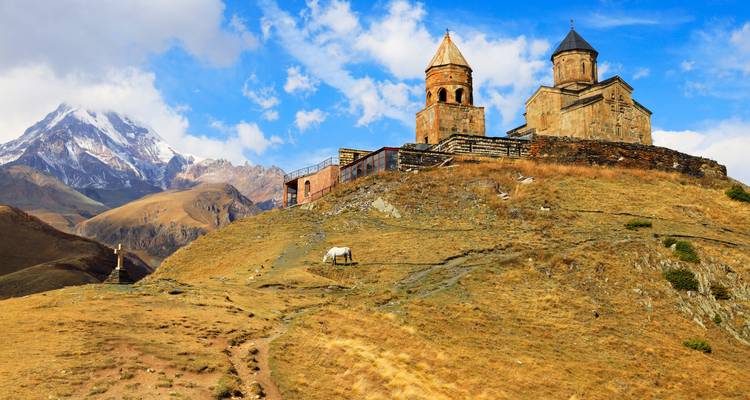 Gergeti-Dreifaltigkeitskirche auf grasigem Bergrücken mit dramatischem schneebedecktem Berg Kazbek im Hintergrund.