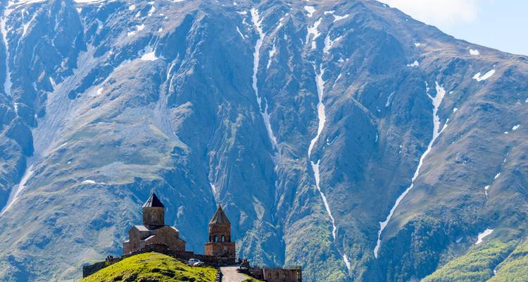 Kleine Kirche auf grünem Hügel vor der aufragenden Felswand des Berges Kazbek mit rauer Landschaft.