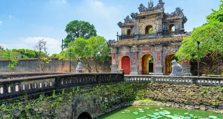 Alter Eingang, umgeben von einem Wassergraben mit Statuen und üppigem Grün.