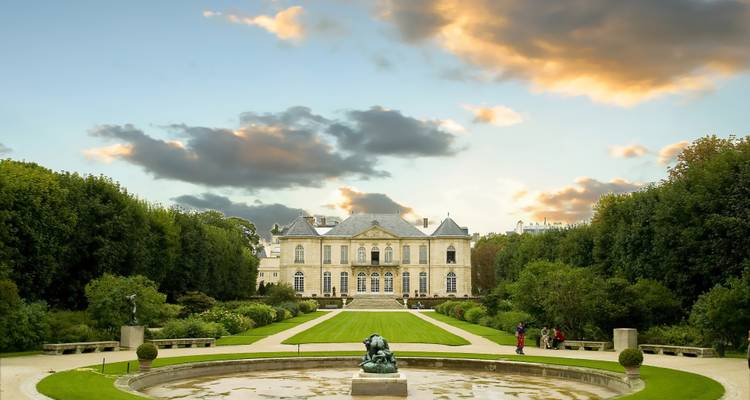 Formaler Garten, der zu einer eleganten Villa mit Wasserbecken und dramatischen Sonnenuntergangswolken führt.