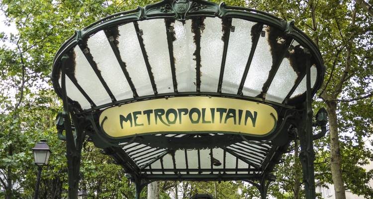 Jugendstil-Eingangsdach aus Schmiedeeisen der Pariser Métro mit „METROPOLITAIN"-Schild unter belaubten Bäumen.