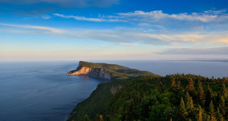 Panoramablick auf die Küste mit bewaldeten Klippen, die in ein ruhiges blaues Meer hineinragen unter einem klaren Abendhimmel.