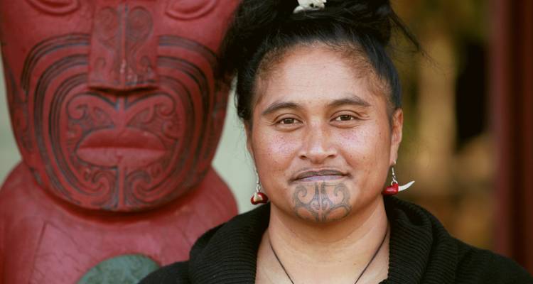 Maori woman with a tattoo on her chin, standing beside a carved figure.