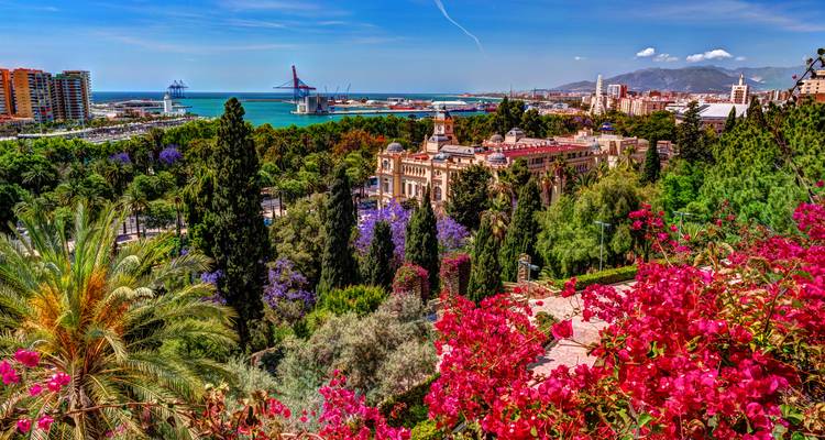 Farbenfroher Ausblick auf Malaga mit blühenden Blumen, dem Hafen, dem Meer und den Stadtgebäuden unter einem klaren Himmel
