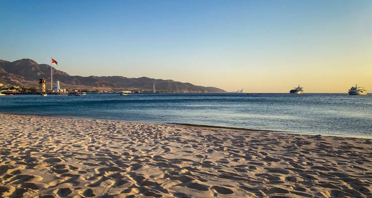 Sandstrand im späten Nachmittagslicht mit jordanischer Flagge und vor Anker liegenden Schiffen in der Ferne.