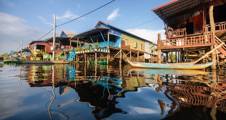 Bunte Stelzenhäuser und Holzboote spiegeln sich in ruhigem Wasser unter klarem blauem Himmel wider.