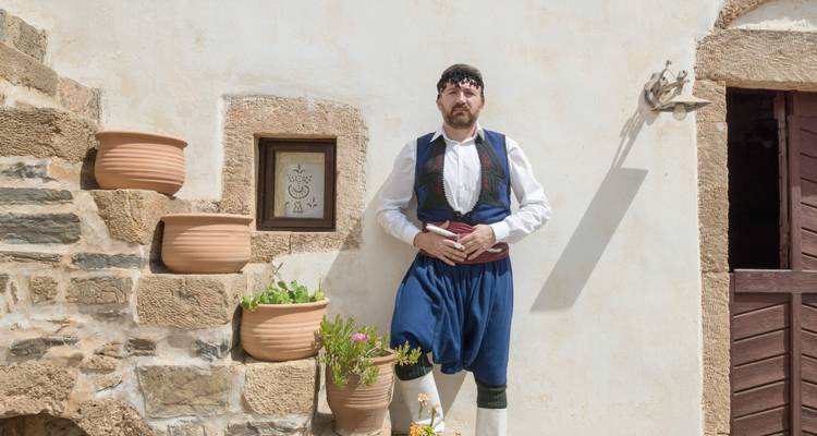 Ein Mann in traditioneller griechischer Kleidung, der vor einer rustikalen Wand steht.