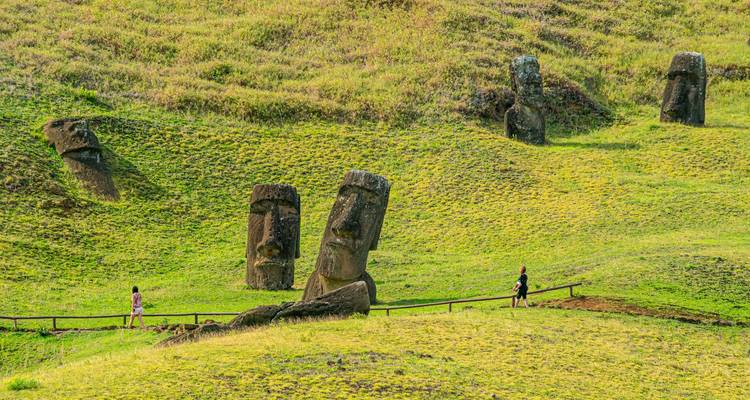 Green rolling hillside dotted with iconic Easter Island Moai statues while two visitors walk along a wooden path.