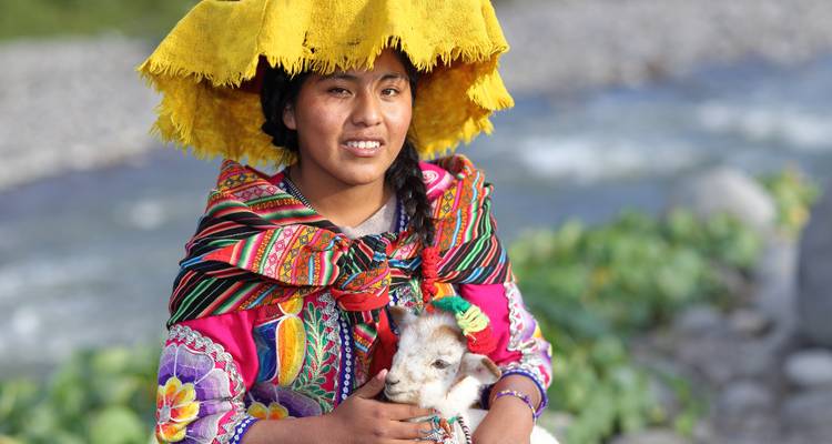 Portrait d'une jeune femme péruvienne en tenue traditionnelle colorée tenant une petite chèvre au bord d'une rivière.