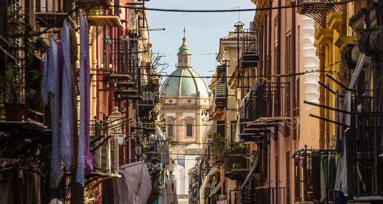 Ruelle sicilienne étroite encadrée par des volets colorés et du linge étendu avec une église à dôme qui s'élève à l'extrémité.