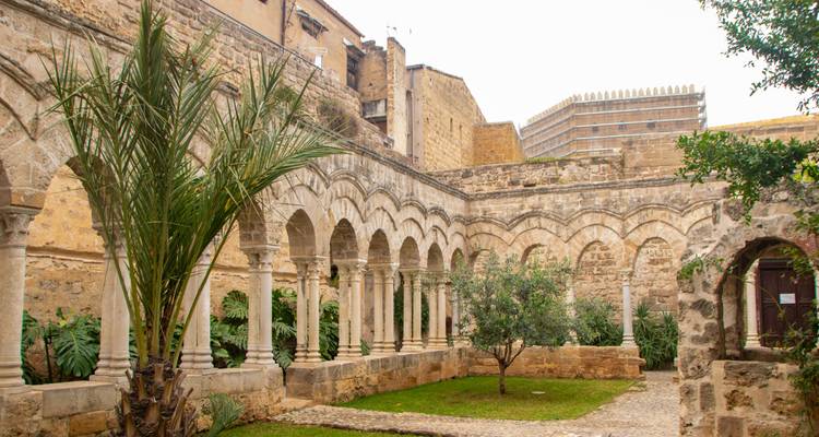 Jardin de cloître paisible entouré d'arcades en pierre et de grands palmiers à l'intérieur d'un monastère historique sicilien.