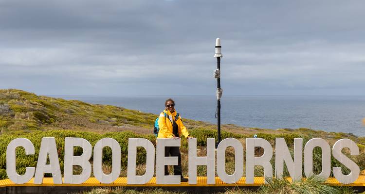 Reiziger in gele jas staande achter grote witte letters die CABO DE HORNOS spellen met zee als achtergrond