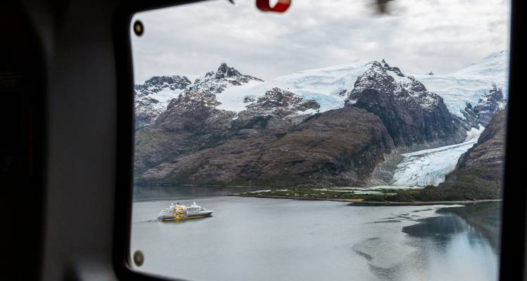 Uitzicht door een raam van een cruiseschip dat voor anker ligt onder sneeuwbedekte pieken en een gletsjer