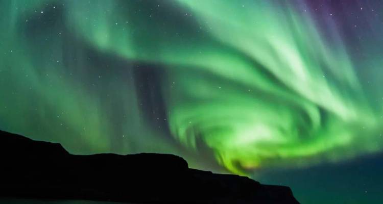 Aurora boreal de un verde vibrante girando a través de un cielo nocturno estrellado sobre acantilados costeros oscuros.