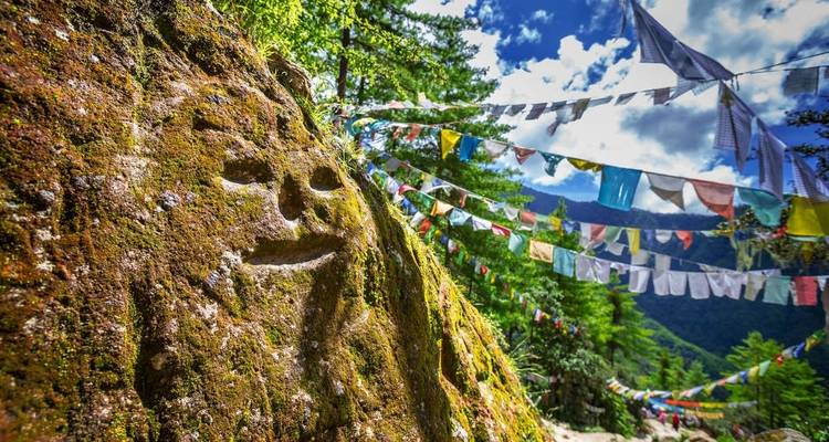 A carved rock face with colorful prayer flags hanging above in a forested area.