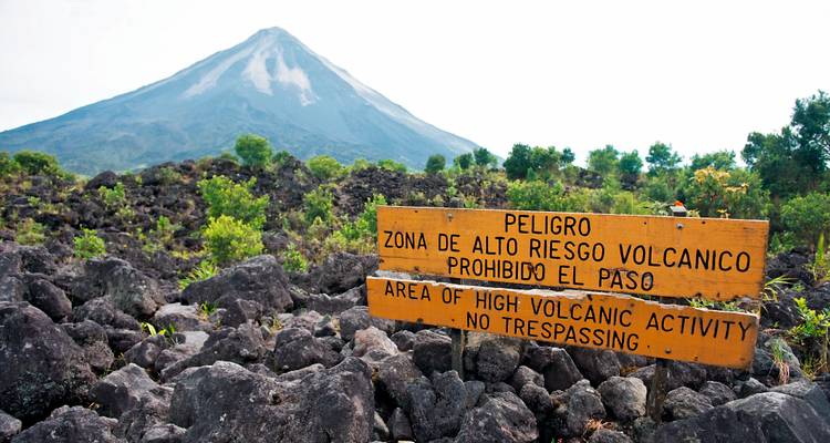 Panneau d'avertissement concernant le risque volcanique élevé installé devant le cône imposant du volcan Arenal et les roches de lave noire.