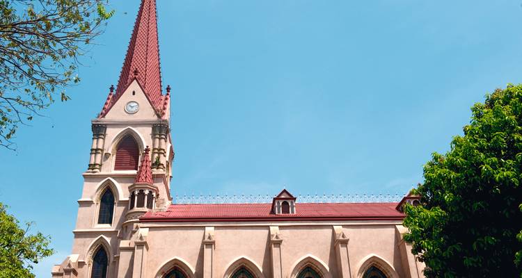 Église élancée de couleur beige aux flèches imposantes, dotée d'un toit rouge, se dressant sur un ciel turquoise limpide et encadrée par des arbres.