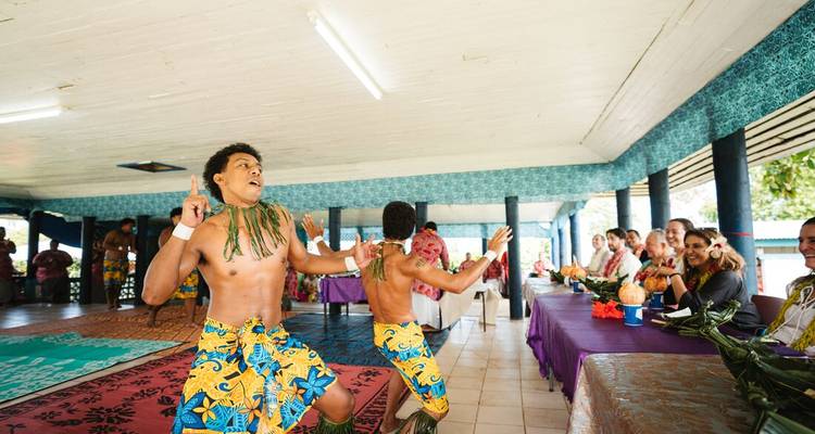 Samoan dancers in bright costumes perform an energetic routine before an audience seated at banquet tables.