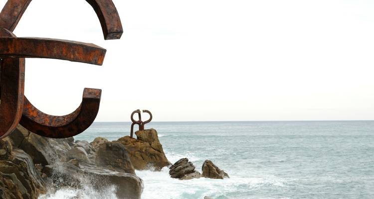 Esculturas de hierro oxidado junto al mar, colocadas sobre formaciones rocosas, con olas rompiendo.