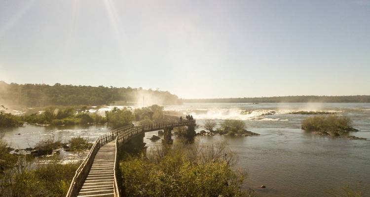 Une passerelle en bois s'étend vers les chutes brumeuses d'Iguazu sous un ensoleillement éclatant.