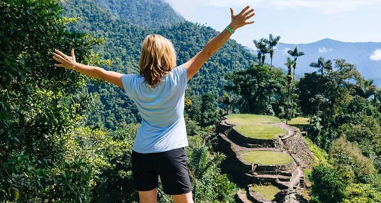 Een wandelaar steekt triomfantelijk zijn armen in de lucht terwijl hij uitkijkt over de stenen terrassen van de Verloren Stad temidden van de dichte jungle.