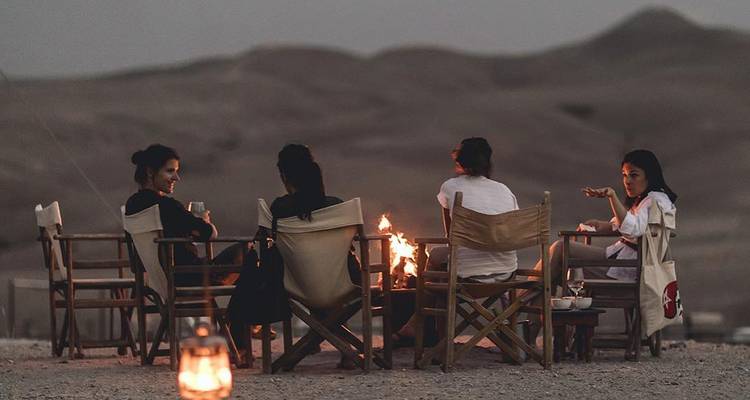 Cinq femmes sont assises sur des chaises pliantes autour d'un petit feu de camp dans un paysage désertique aride au crépuscule, avec de douces montagnes se découpant en silhouette en arrière-plan.