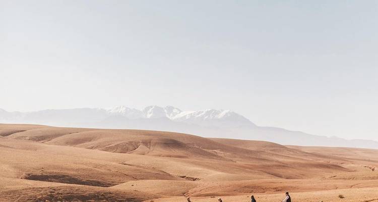 Vaste plaine désertique avec de minuscules randonneurs au premier plan et les montagnes de l'Atlas enneigées sous un ciel pâle.