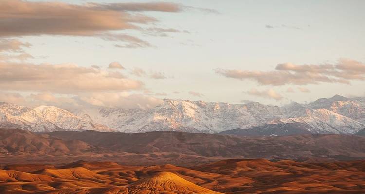 La lumière dorée baigne la chaîne de montagnes lointaine couverte de neige au-dessus des crêtes désertiques ambrées sous des nuages épars.