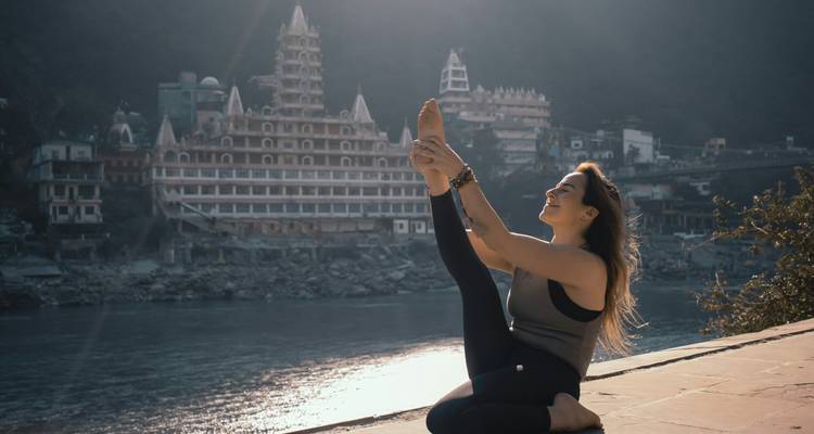 Young woman practicing yoga on a riverside platform with a multi-tiered temple across the water.