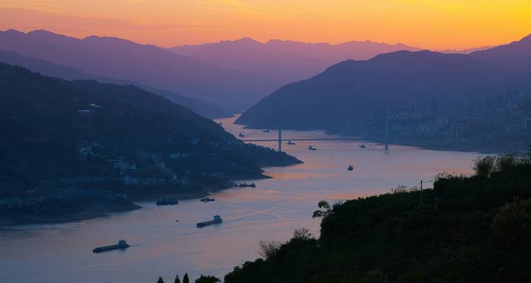 Sunset over a winding river gorge dotted with cargo barges and layered mountain silhouettes.
