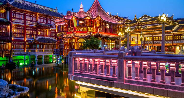 Illuminated traditional Chinese pavilions and bridges reflect in a pond at blue hour.