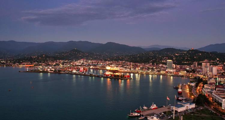 Avondbeeld van een verlichte stedelijke skyline met een waterkant en bergen.