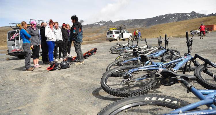 Travellers gather beside a van and rows of mountain bikes on a barren high-altitude plain dwarfed by snowy peaks.