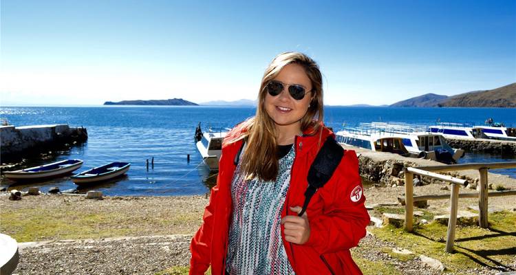 Young woman in red jacket smiles with moored boats and shimmering lake under clear skies.