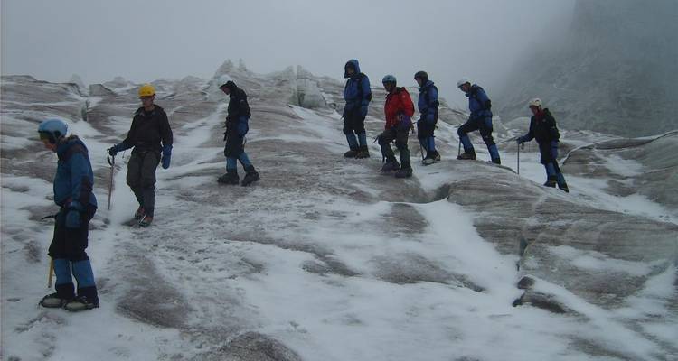 Line of trekkers with ice axes walks across a misty, snow-covered glacier slope.