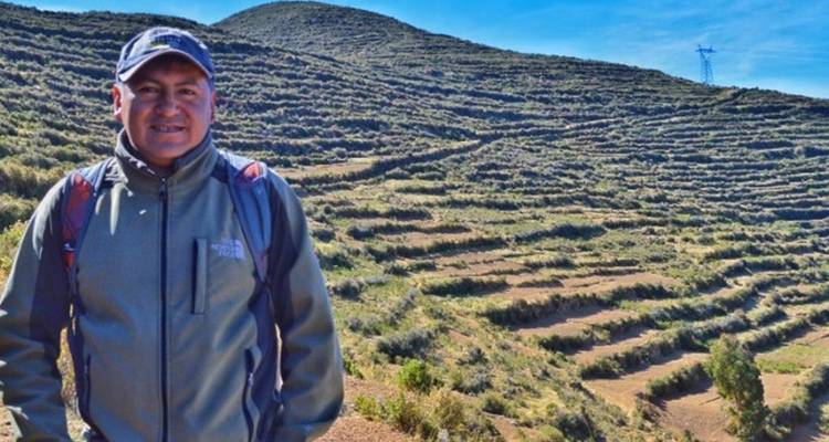 Un homme avec un sac à dos pose à côté de terrasses agricoles anciennes sous un ciel andin dégagé.