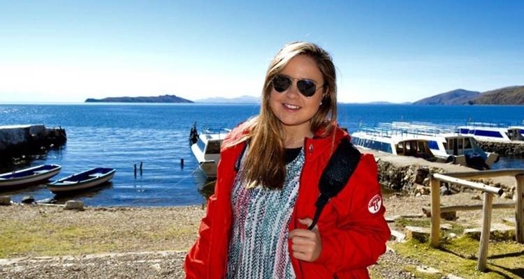 Un voyageur vêtu d'une veste rouge se tient sur une jetée au bord d'un lac avec des bateaux et une eau bleu profond en arrière-plan.
