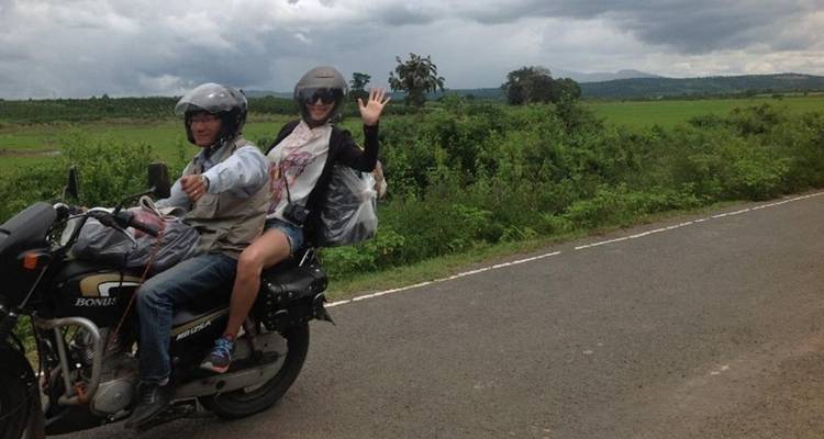 Deux voyageurs portant des casques saluent joyeusement en chevauchant une motocyclette le long d'une route rurale bordée de champs verdoyants et luxuriants et de collines lointaines sous un ciel menaçant.