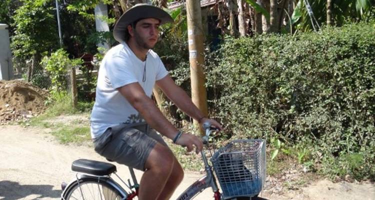 Un jeune homme portant un chapeau à large bord fait du vélo sur une route poussiéreuse de village bordée d'arbustes, d'arbres et de maisons rustiques.