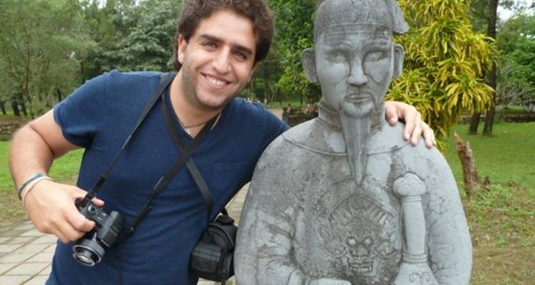 Un touriste souriant muni d'un appareil photo passe un bras autour d'une statue de guerrier en pierre grise située dans un jardin historique verdoyant.